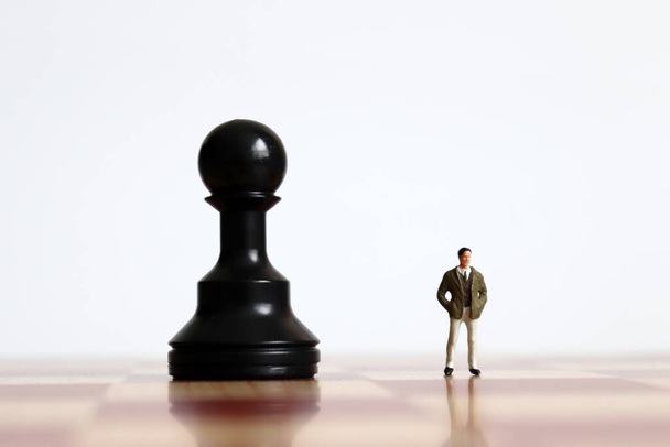 Un hombre en miniatura de pie lado a lado con un peón negro en un tablero de ajedrez
. - Foto, Imagen