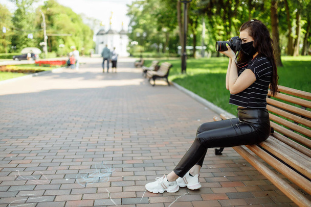 mooi meisje met een camera draagt een medisch masker en maakt foto 's in het park. Coronavirus 2019 nCoV pneumonie uitbraak. - Foto, afbeelding