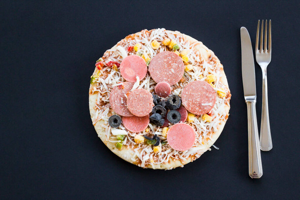 Άψητη κατεψυγμένη στρογγυλή πίτσα σε μαύρο χρώμα με πιρούνι και μαχαίρι. - Φωτογραφία, εικόνα