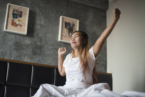 Happy asian young woman stretching after waking up while in the bedroom - Фото, изображение
