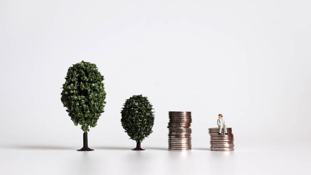 Dwa drzewa obok siebie z miniaturowym mężczyzną siedzącym na stosie monet. - Zdjęcie, obraz
