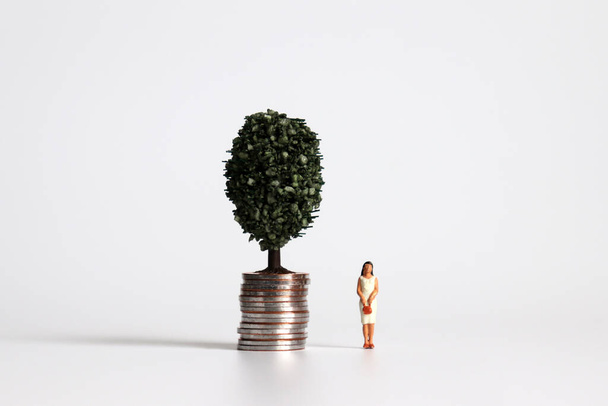 Μια γυναίκα μινιατούρα στέκεται δίπλα σε ένα σωρό από νομίσματα με δέντρα. - Φωτογραφία, εικόνα