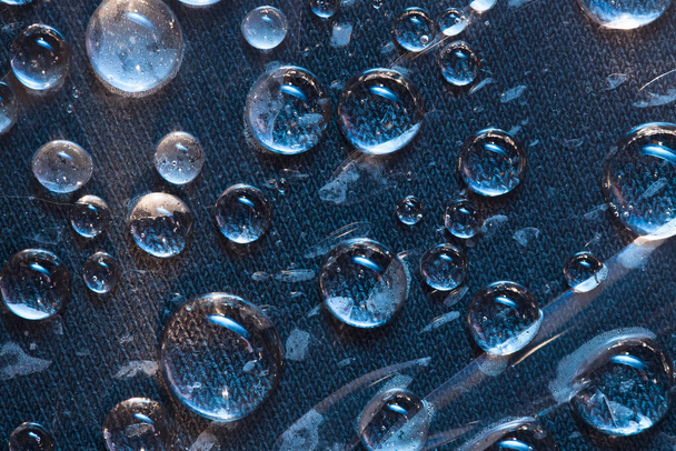 Gotas azules transparentes de agua son iluminadas por el sol brillante. Fondo de tela azul. De cerca.
. - Foto, Imagen
