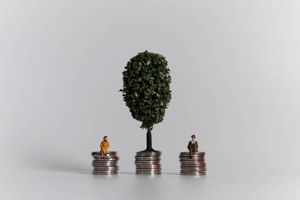 Minyatür bir kadın ve erkek aynı yükseklikte bozuk paraların üzerinde oturuyorlar. Kadın ve erkek için maaş ve terfi eşitliği kavramı. - Fotoğraf, Görsel