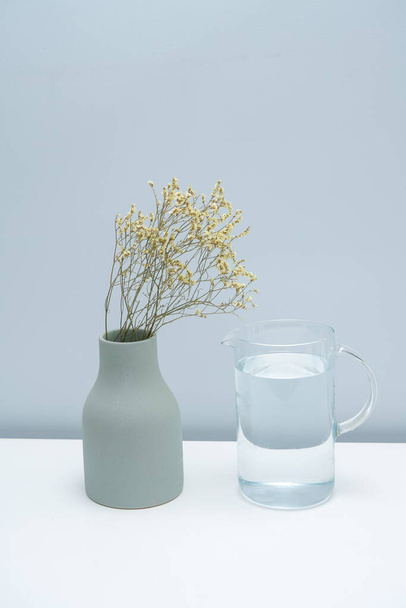 Um vaso com flores secas e um copo de água na mesa branca
. - Foto, Imagem