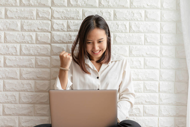 Asian young woman sitting in living room smile celebrating and using computer laptop for using social media and shopping online feeling so happiness and cheerful - 写真・画像