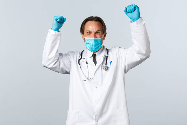 Covid-19, preventing virus, healthcare workers and vaccination concept. Happy excited doctor in medical mask and gloves raise hands up shouting yes, winning, celebrating victory or achievement - Fotó, kép