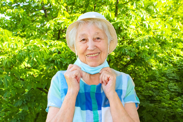 積極的な笑顔シニア女性は屋外から保護医療マスクを離陸します.幸せな終わりの概念。花粉症。コロナウイルスの勝利だ。Pandemic Covid-19.夏の緑の木 - 写真・画像