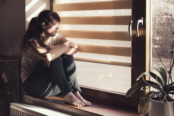 Traurige junge Frau, die zu Hause isoliert am Fenster sitzt und aufpasst. Coronavirus-Quarantäne-Konzept. - Foto, Bild