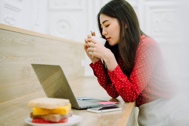 Asyalı, uzun, siyah saçlı, kırmızı tişörtlü güzel bir kadın dizüstü bilgisayarın önünde beyaz bardaktan sıcak kahve içiyor. Evde çalışıyorum. Evden çalış.. - Fotoğraf, Görsel