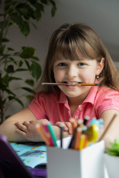Menina estudante senta-se em uma mesa e segura um lápis em sua boca. Orientação vertical. Aprendizagem divertida caseira
 - Foto, Imagem