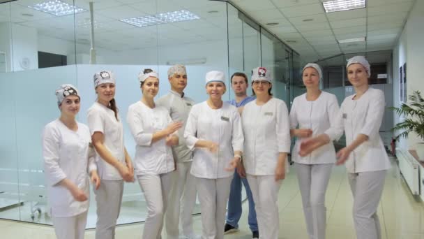 Μια ομάδα επαγγελματιών ιατρών με άσπρη ιατρική στολή διπλώνουν τα χέρια τους στο λόμπι μιας σύγχρονης κλινικής και χαμογελούν. - Πλάνα, βίντεο