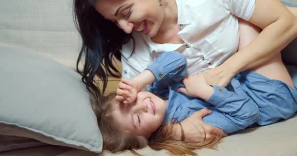 Cheerful family mother and daughter embracing and laughing lying on bed together. Happy young mom tickling her cute little daughter enjoying communication at home. - Filmagem, Vídeo