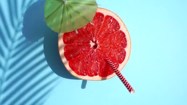 Grapefruit (oranžový) rotující s koktejlovým deštníkem ve slunečním světle na barevném modrém pozadí. Ovocné šťávy, relaxační, tropické a relaxační koncepce. - Záběry, video