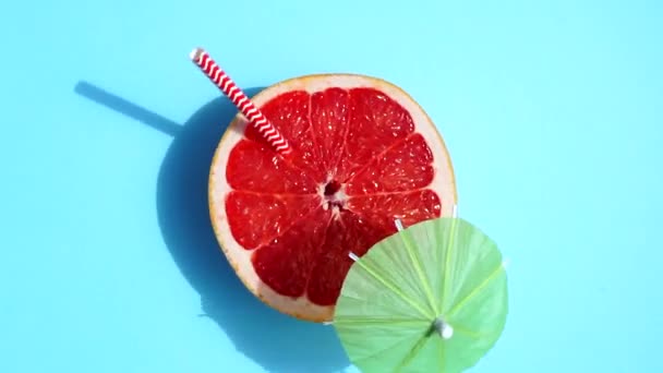Pompelmo (arancio) ruotante con ombrellone da cocktail alla luce del sole su fondo blu colorato. Succhi di frutta, relax, tropici e relax. - Filmati, video
