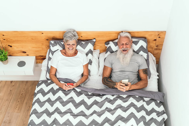 Heureux couple de personnes âgées utilisant des smartphones mobiles ci-dessous couvertures au lit - Les personnes âgées ayant le temps de coucher drôle regarder sur les tendances des nouveaux médias - Style de vie de relation amoureuse des personnes âgées et concept de technologie - Photo, image