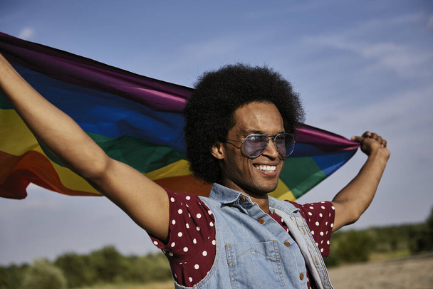 Junger Afrikaner mit Regenbogenfahne steht am Strand  - Foto, Bild