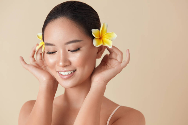 Face. Woman With Flowers Portrait. Beautiful Asian Model With Plumeria In Hair Posing Against Beige Background. Charming Girl With Closed Eyes, Nude Makeup, Healthy, Hydrated, Smooth And Glowing Skin. - Foto, Imagen
