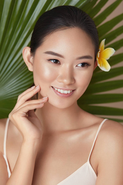 Beauty. Woman On Leaf Background Portrait. Beautiful Asian Model With Flower In Hair Touching Face And Looking Away. Young Female With Perfect Skin And Natural Makeup After Using Organic Cosmetic.  - Fotografie, Obrázek