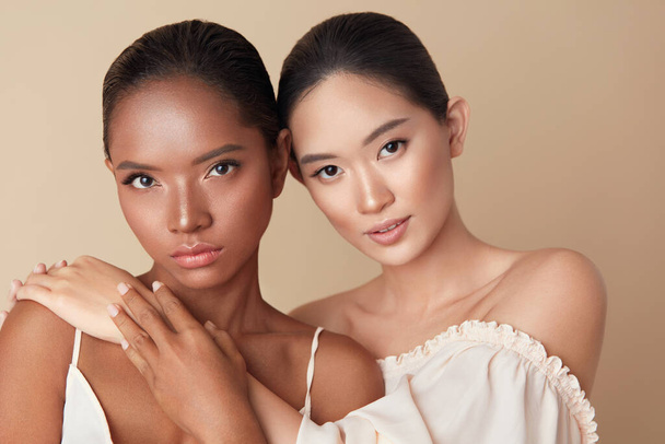 Diverse. Women Beauty Portrait. Multi-Ethnic Models With Natural Makeup And Perfect Skin Against Beige Background. Mixed Race And Asian Girls Standing Together, Hug Each Other And Looking At Camera. - 写真・画像