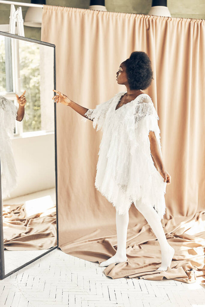 full-length photo of a dark-skinned girl in a white boho style dress, white tights, on a brown fabric background, she looks in the mirror - Foto, afbeelding