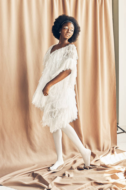full-length photo of a dark-skinned girl in a white boho style dress, white tights, on a brown fabric background she looks to the right side holding the dress  - Foto, imagen