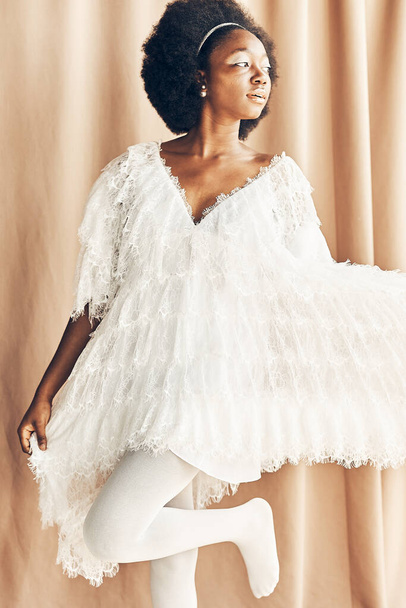 full-length photo of a dark-skinned girl in a white  boho style dress, white tights, on a brown fabric background she looks to the right side holding the dress  - Foto, afbeelding