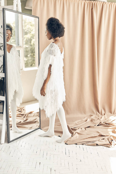 полноразмерное фото темнокожей девушки в белом платье в стиле бохо, белые колготки, на коричневом фоне, она смотрит в зеркало
 - Фото, изображение