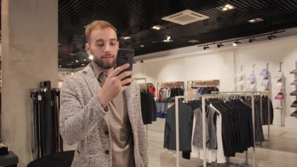 Elegante hombre de negocios guapo en un traje y barba hablando en videollamada usando un teléfono inteligente en una tienda de ropa para hombres
. - Imágenes, Vídeo