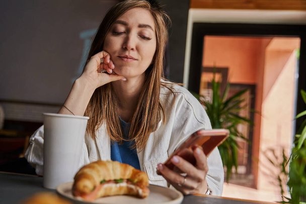 Vooraanzicht van een aantrekkelijke vrouw die een bril draagt met een smartphone, koffie drinkt in een café. Vrouwelijke sms 'en en berichten delen op sociale media, genieten van mobiele technologie, ontspannen in de coffeeshop. - Foto, afbeelding