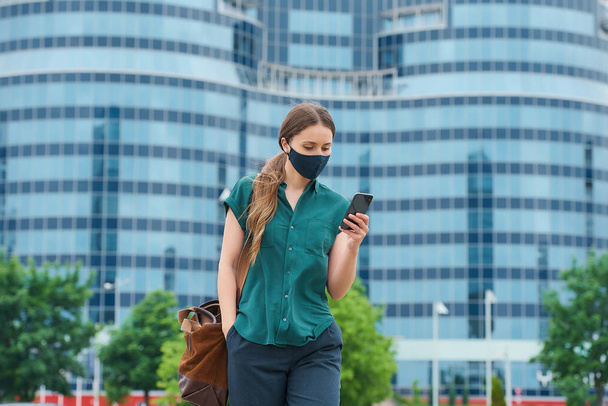 Hezká mladá žena v námořně modré lékařské masce obličeje, aby se zabránilo šíření koronaviru procházky v centru města. Dívka s dlouhými vlasy čte zprávy na smartphonu v centru města. - Fotografie, Obrázek