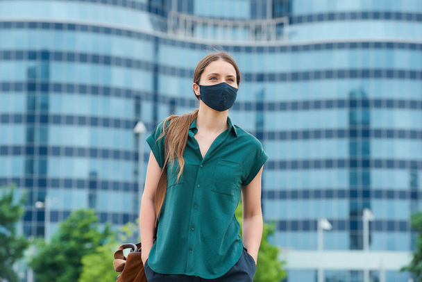 Μια όμορφη νεαρή γυναίκα με μάσκα ιατρικού προσώπου για να αποφύγει την εξάπλωση του coronavirus περπατώντας στο κέντρο της πόλης. Ένα κορίτσι με μακριά μαλλιά που κρατάει κοινωνική απόσταση φοράει προστατευτική μάσκα στο κέντρο της πόλης - Φωτογραφία, εικόνα