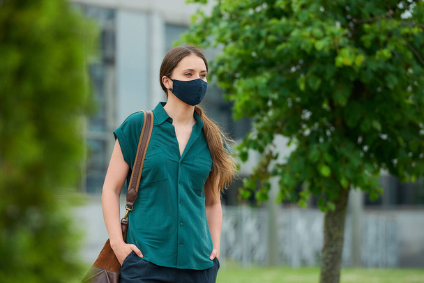 Woman in medical face mask walks between trees thrusting hand into pocket of trousers, holds a bag in downtown. Girl keeping social distance wears a protective mask to avoid the spread of coronavirus - Photo, Image