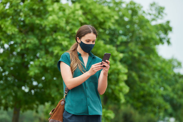 Nuori nainen laivaston sinisessä lääketieteellisessä kasvonaamiossa lukee uutisia älypuhelimella kävellessään puistossa. Tyttö suojelevassa naamiossa pitää sosiaalista etäisyyttä välttääkseen koronaviruksen leviämisen puiden lähellä.. - Valokuva, kuva