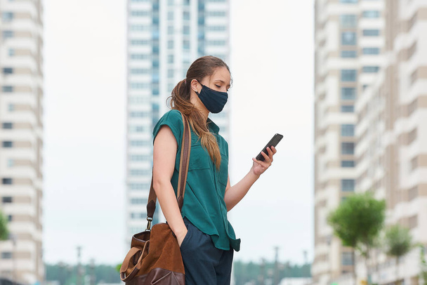 Side view of woman in a medical face mask who reads news on a smartphone thrusting a hand into a pocket of trousers walks between skyscrapers. Girl keeping social distance wearing protective face mask - Foto, Imagem