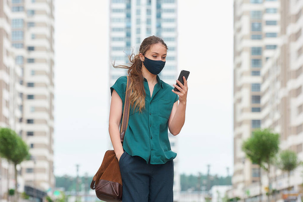 A young woman in a medical face mask reads news on her smartphone thrusting a hand into a pocket of trousers walks between skyscrapers. A girl keeping social distance wearing a protective face mask. - Fotografie, Obrázek