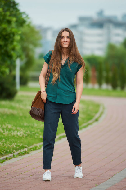Nuori nainen pukeutuu tyylikäs rento vaatteita nauttia hänen kävellä puistossa. Tyttö pitkät hiukset kuluu toimisto tyyli vaatteita pitää sosiaalisen etäisyyden kaupungissa. - Valokuva, kuva