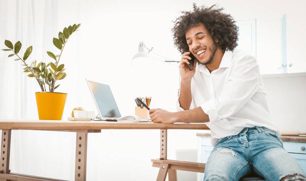 Side view of young entrepreneur or student talking mobile phone while working from home. Charming Arab guy chatting on smart phone with friends or customers. Tinted image - Фото, зображення