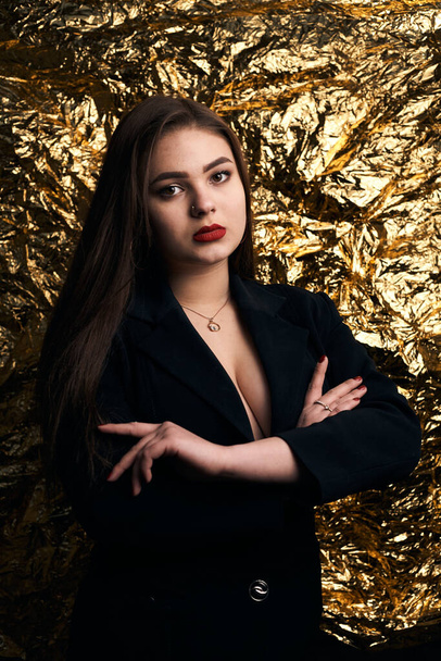Stilvolles molliges Mädchen posiert in einer schwarzen Jacke auf goldenem Hintergrund. - Foto, Bild