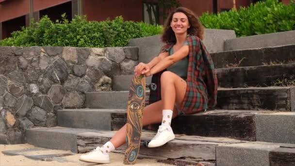 Schöne kaukasische tätowierte Frau in Jeanshosen, kariertem Hemd sitzt bei Sonnenuntergang auf der Treppe und hält Longboard, Skateboard   - Filmmaterial, Video