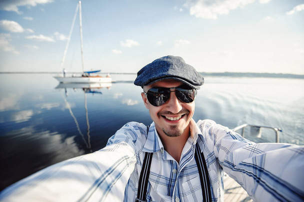 A successful businessman and yachtman  resting on his yacht. Good life. Beautiful bearded man wearing sunglasses and hat, doing sephi on the background of a river and yacht. - Foto, immagini