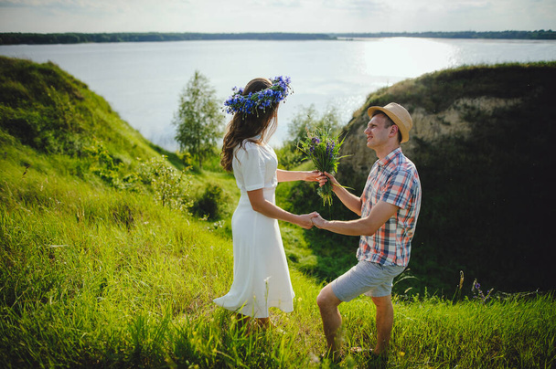 Milující mladý muž dává kytici divokých květin své přítelkyni. Krásná dívka v věnec a stylový chlap v klobouku tráví čas venku. Milostný příběh. - Fotografie, Obrázek