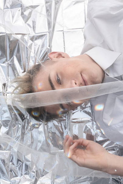 A handsome model guy lies under a tulle fabric on silver foil. - Foto, Imagen