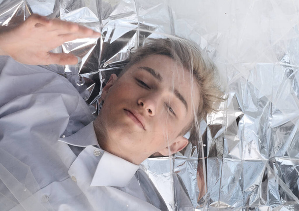 A handsome model guy lies under a tulle fabric on silver foil. - Photo, Image
