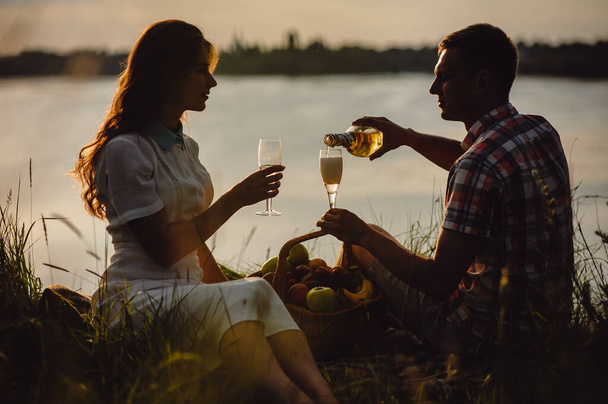 Siluetti rakastava pari juo viiniä auringonlaskun aikaan. Mies ja tyttö romanttisilla treffeillä, istuu hedelmäkorin lähellä ja juo samppanjaa laseista. - Valokuva, kuva