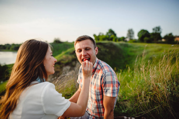 Smiling portrait of a man whom a woman feeds strawberries outdoors. Loving couple boyfriend and girlfriend having picnic on river bank. - Foto, Imagem