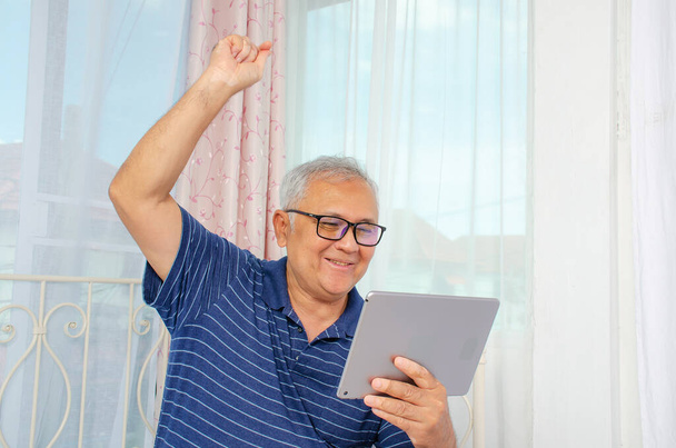 Hombre mayor sentado en el sofá se sienten felices y encantadores cuando se mira a la tableta. Hombre mayor levantando la mano y gritando, concepto de jubilación feliz
. - Foto, Imagen