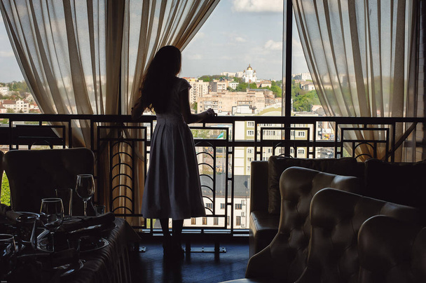 Slim barna, fehér ruhában nézi a várost ablakból az étteremben - Fotó, kép