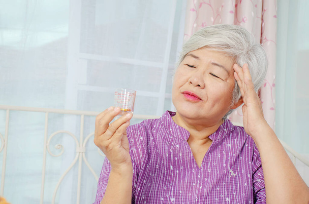 Yaşlı bir kadın cam bardağa bakıyor. İlaçları kontrol ediyor ve başını tutuyor. Şiddetli migren ağrısı çekiyor. - Fotoğraf, Görsel