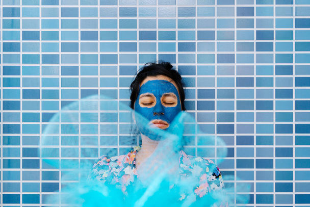Γυναίκα σκέφτεται με τα μάτια κλειστά χρησιμοποιώντας θεραπεία ομορφιάς και διαλογισμό σε μπλε μπάνιο τοίχου, υπάρχει ένα μπάνιο σφουγγάρι μπροστά της - Φωτογραφία, εικόνα